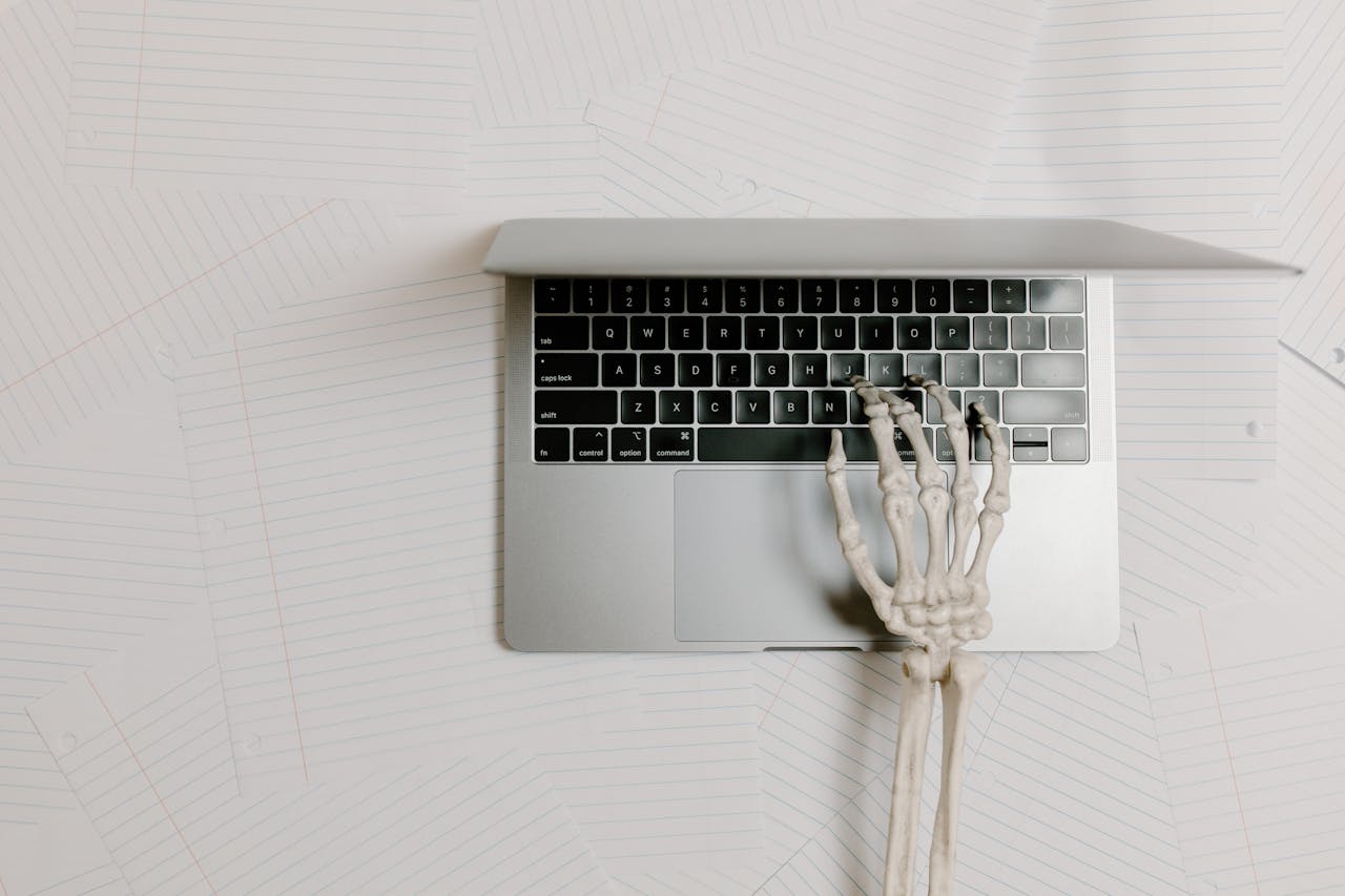 A conceptual overhead view of a skeleton hand typing on a laptop over scattered papers.